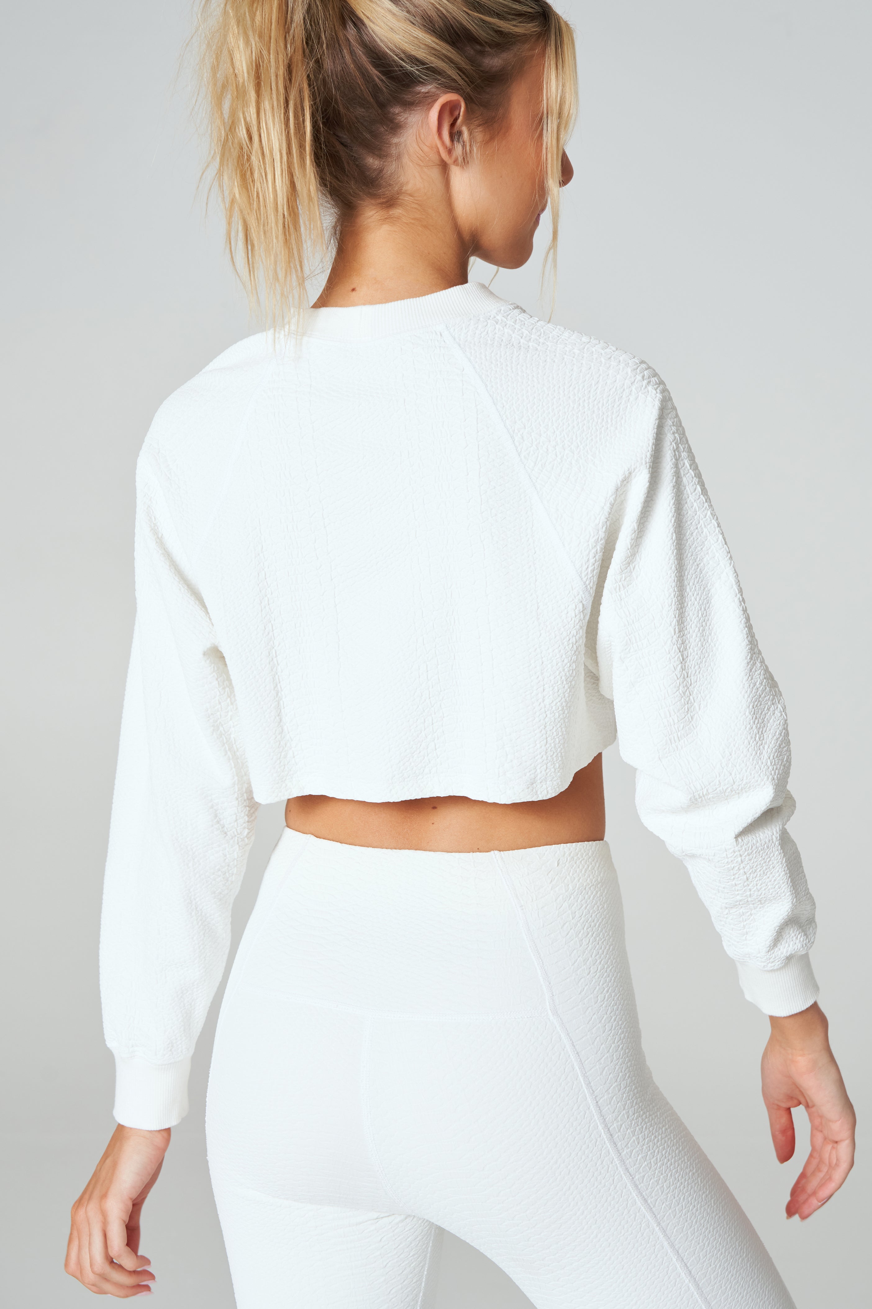 A woman in a textured workout set faces away, with her ponytail swaying. The ensemble includes a ribbed crew neck crop top and high-waisted leggings, set against a plain light gray background that highlights the sleek design resembling the Isabella Pullover by Gigi C Sport.