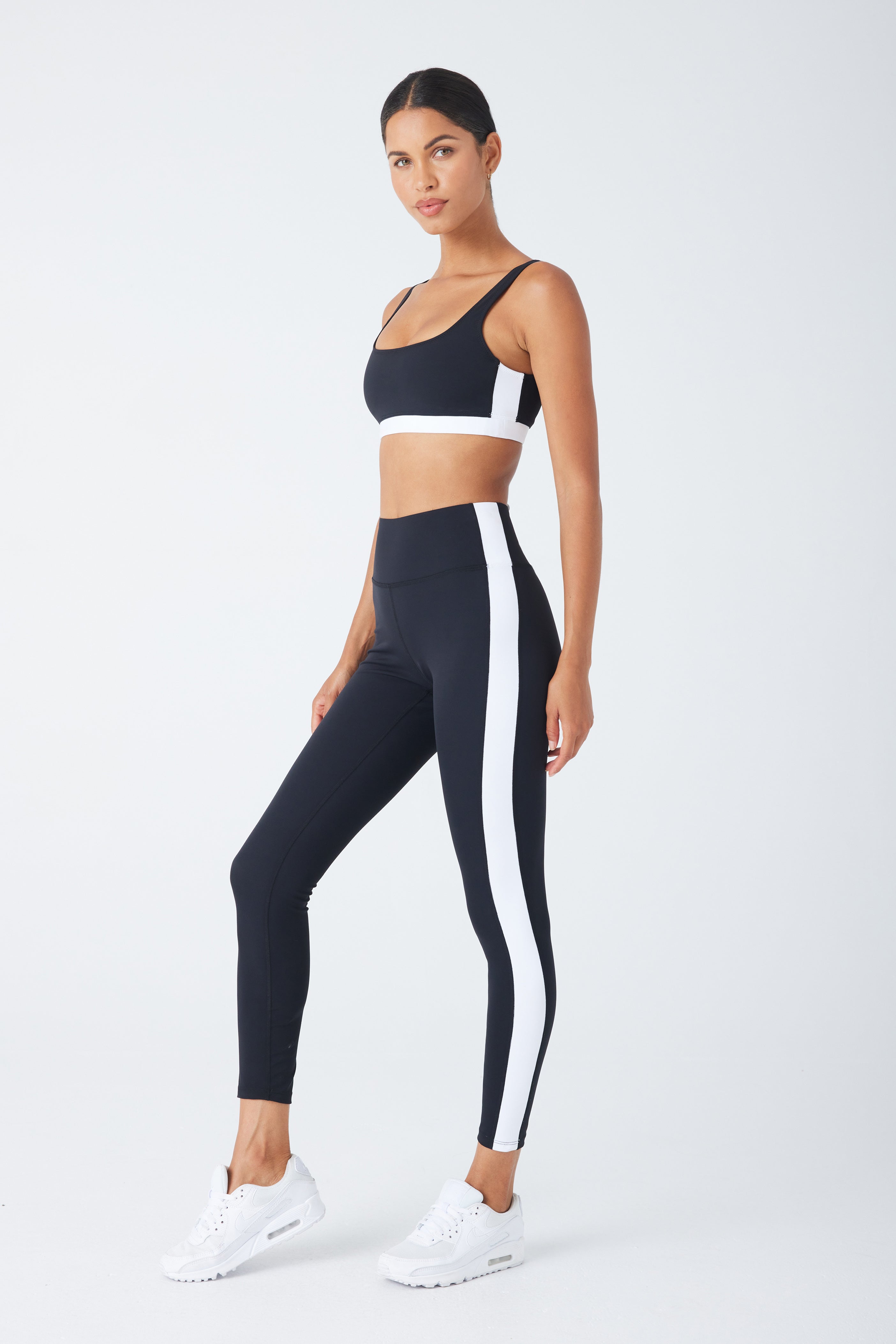 The person is confidently wearing Gigi C Sports Cora Legging, high-waisted and made from recycled black and white jersey, paired with a matching sports bra. They stand against a plain backdrop in white sneakers, hair styled back, with one foot slightly forward.