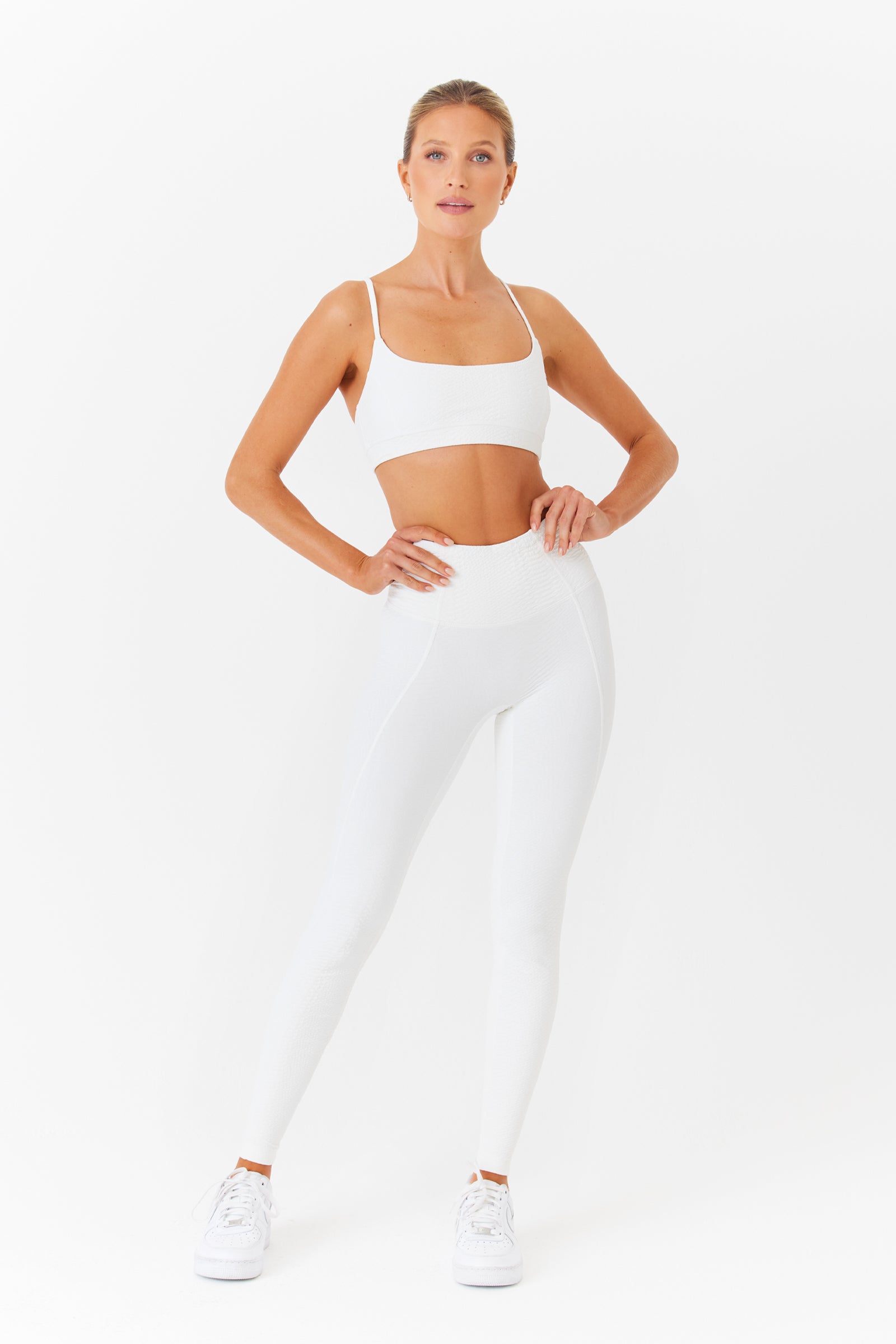 A person confidently stands with hands on hips in a white Gigi C Sport Emery Bra and high-waisted leggings featuring loop strap detail. Paired with white sneakers, the breathable knit design radiates style and comfort against a plain white background.