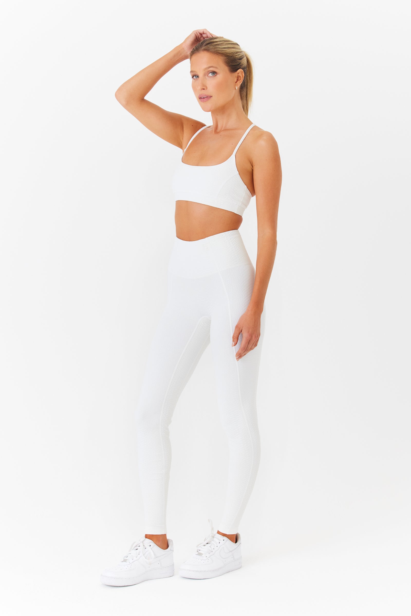 A person poses against a white background, wearing the Emery Bra from Gigi C Sport with high-waisted leggings that have loop strap details. The breathable knit ensemble is paired with white sneakers, and one hand rests on their head.