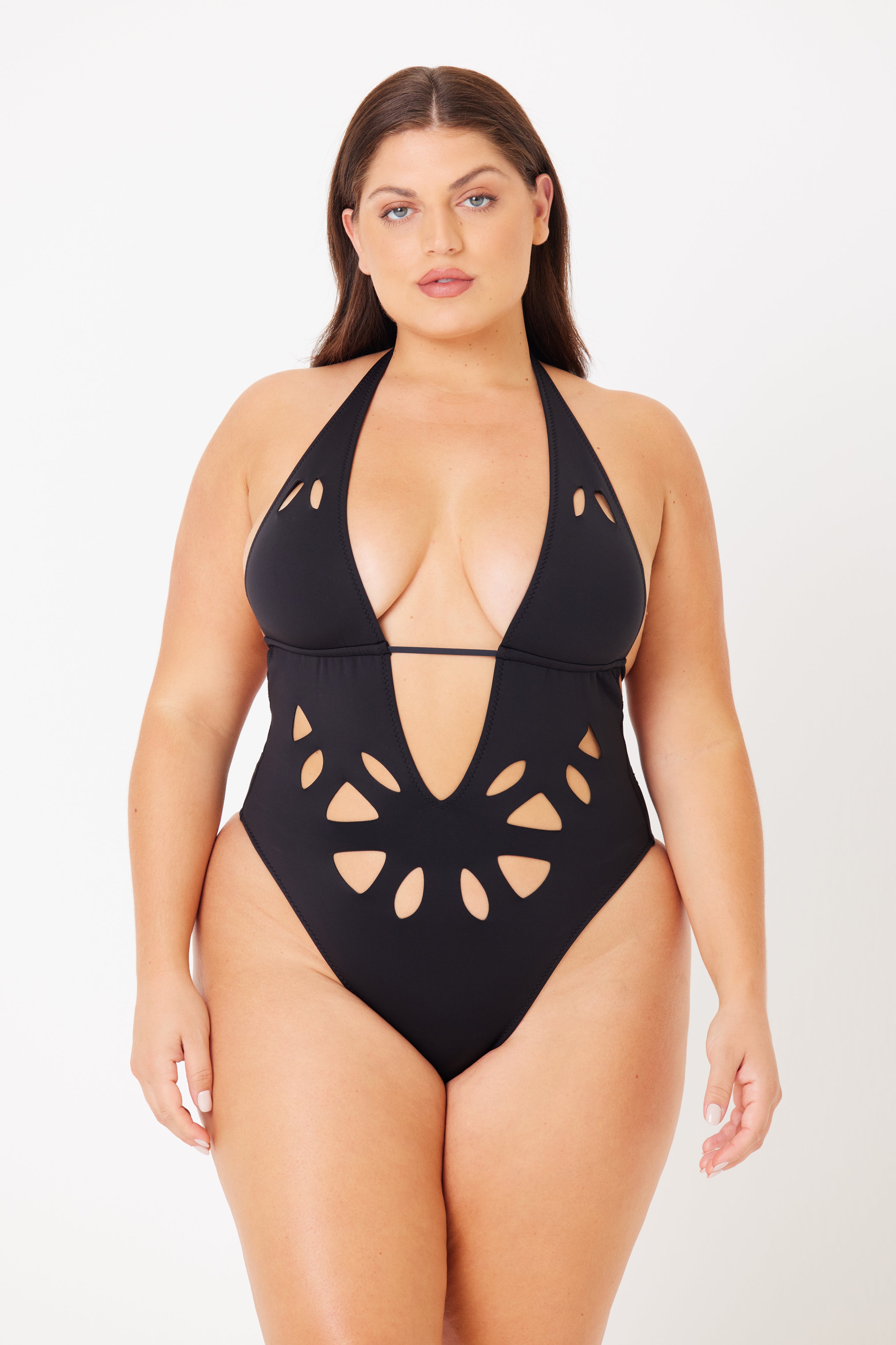 A woman confidently wears the Casey Plunging One-Piece by Gigi C Bikinis, showcasing peekaboo cutouts and a stylish halter neck with a deep neckline, standing against a plain, light background and exuding modern elegance.