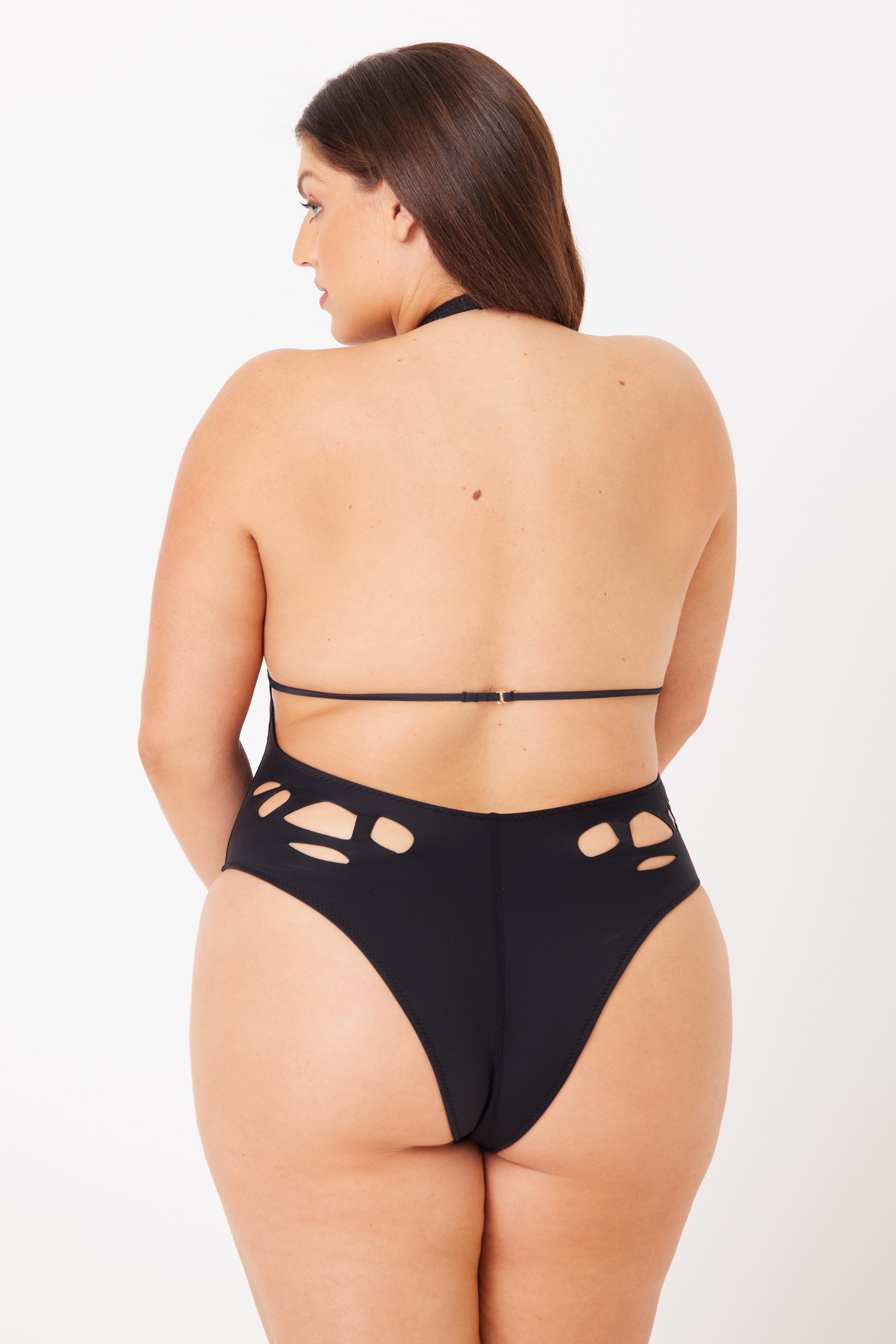 A woman poses with her back to the camera in a Casey Plunging One-Piece by Gigi C Bikinis. The black swimwear, featuring peekaboo cutouts and striking side designs akin to lasercut technology, complements her glance over the shoulder against a plain white backdrop.