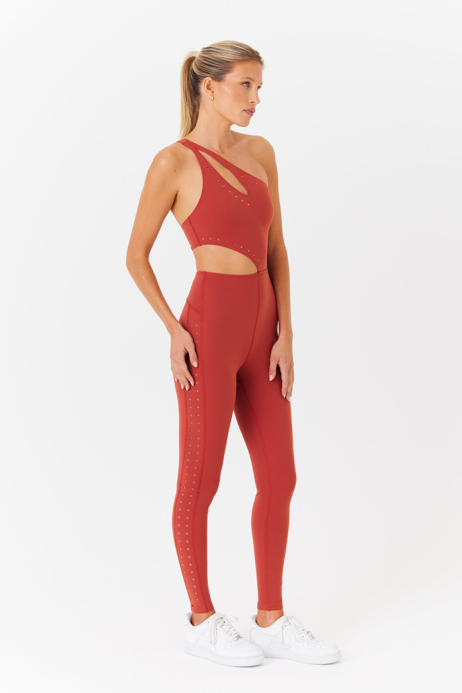 Wearing a red Cassie Jumpsuit by Gigi C Sport with asymmetrical laser cutouts, a person stands sideways against a white background. Their long, light hair is tied back, and theyre sporting white sneakers.