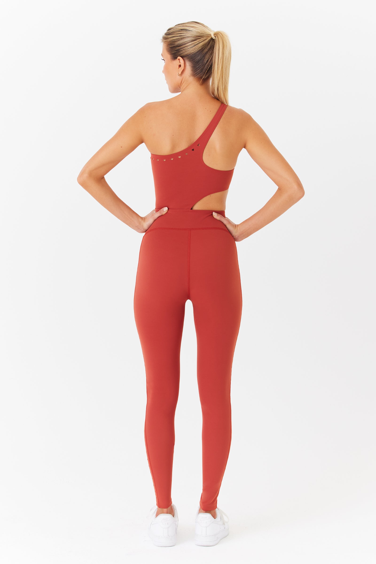 A ponytailed person stands confidently with hands on hips, wearing a rust-colored Gigi C Sport Cassie jumpsuit with laser cutouts on the back, paired with white sneakers. They pose against a plain white background.