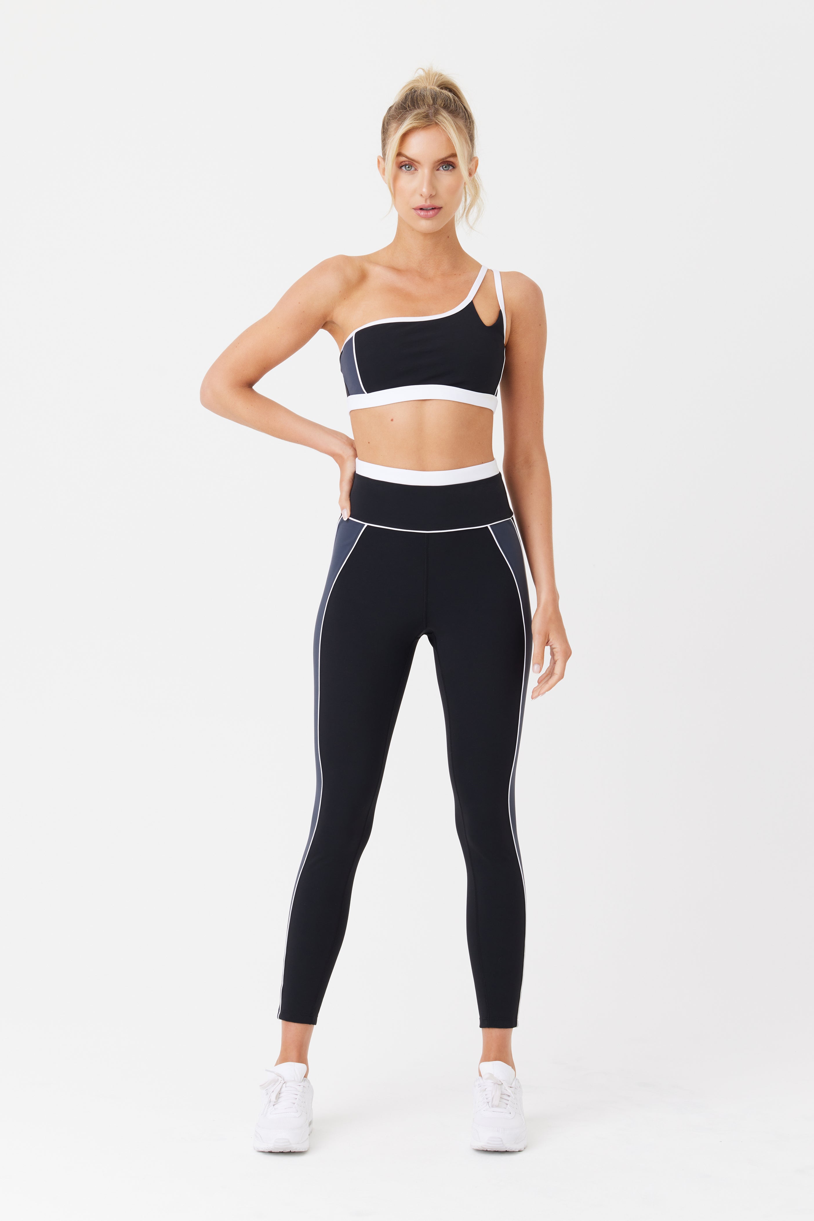 A person confidently poses against a white backdrop in the Gigi C Sport Jocelyn Legging, featuring a black and white one-shoulder athletic top and high-waisted leggings with white piping. The outfit is crafted from breathable recycled polyamide knit, paired with tied-up hair and sleek white shoes.