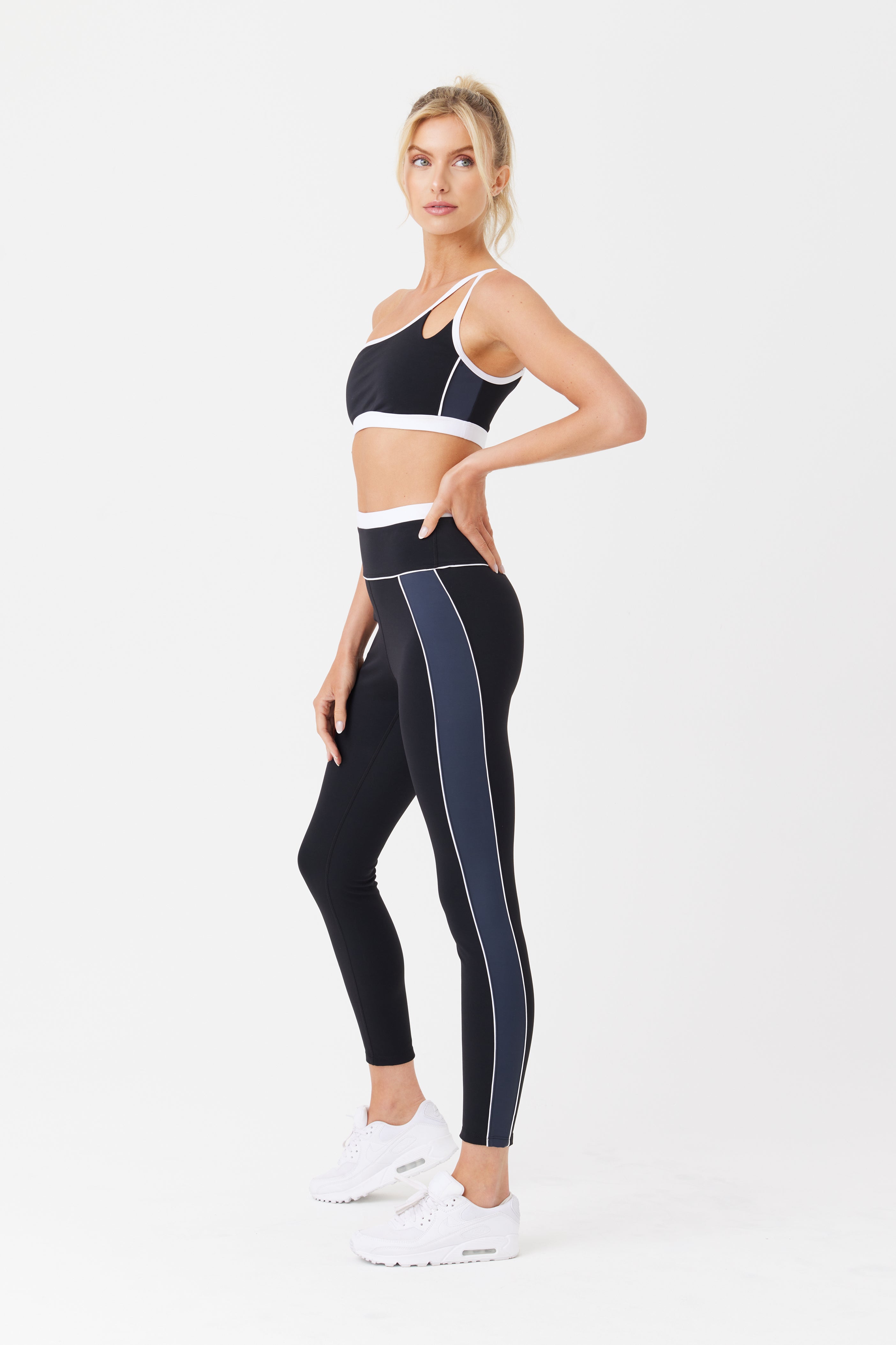 A woman models the Gigi C Sport Jocelyn Legging in black, featuring white piping, against a white backdrop. She teams it with a matching sports bra and white sneakers. Her hair is tied back as she gazes to the side with one hand on her hip, highlighting the outfits breathable knit.