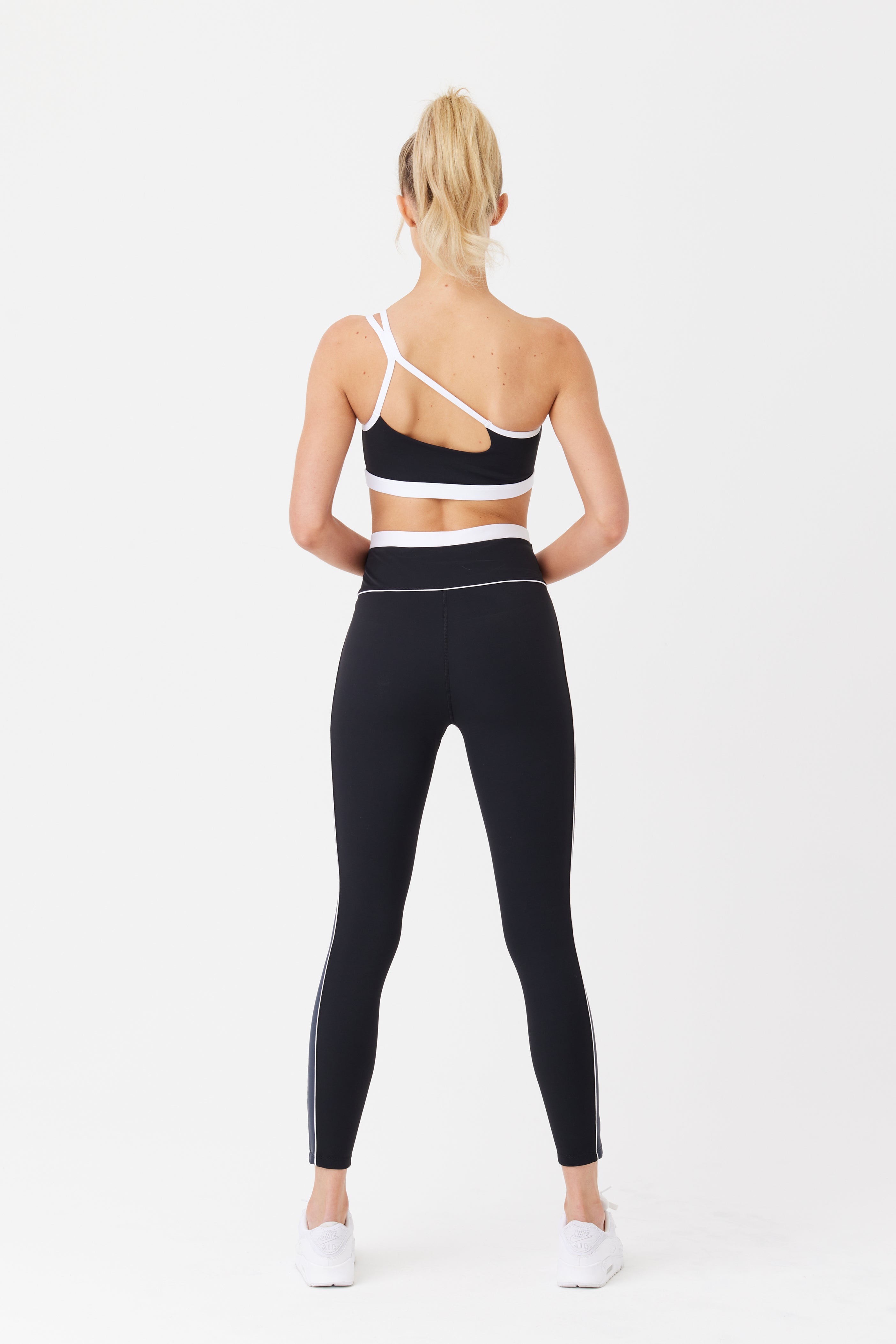 A person in a black and white athletic outfit displays the recycled polyamide one-shoulder sports bra and Gigi C Sports Jocelyn Legging, set against a white backdrop. White sneakers complete the look, with their hair in a high ponytail.