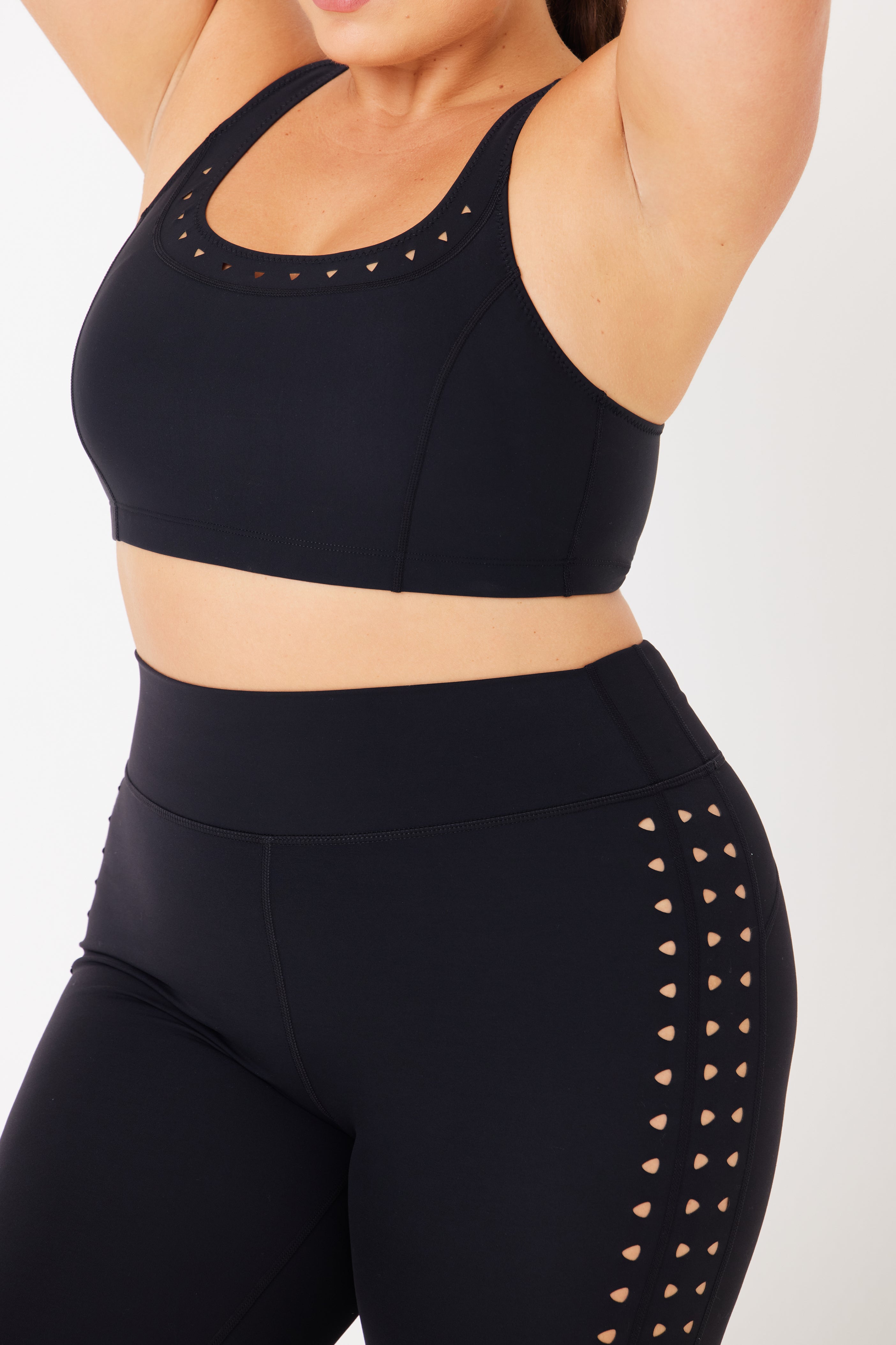 A model wears a Black Annabelle Bra Top and matching leggings from Gigi C Sport, featuring small heart-shaped cutouts on the sides. This form-fitting, breathable knit outfit is perfect for exercise and is set against a plain white background.