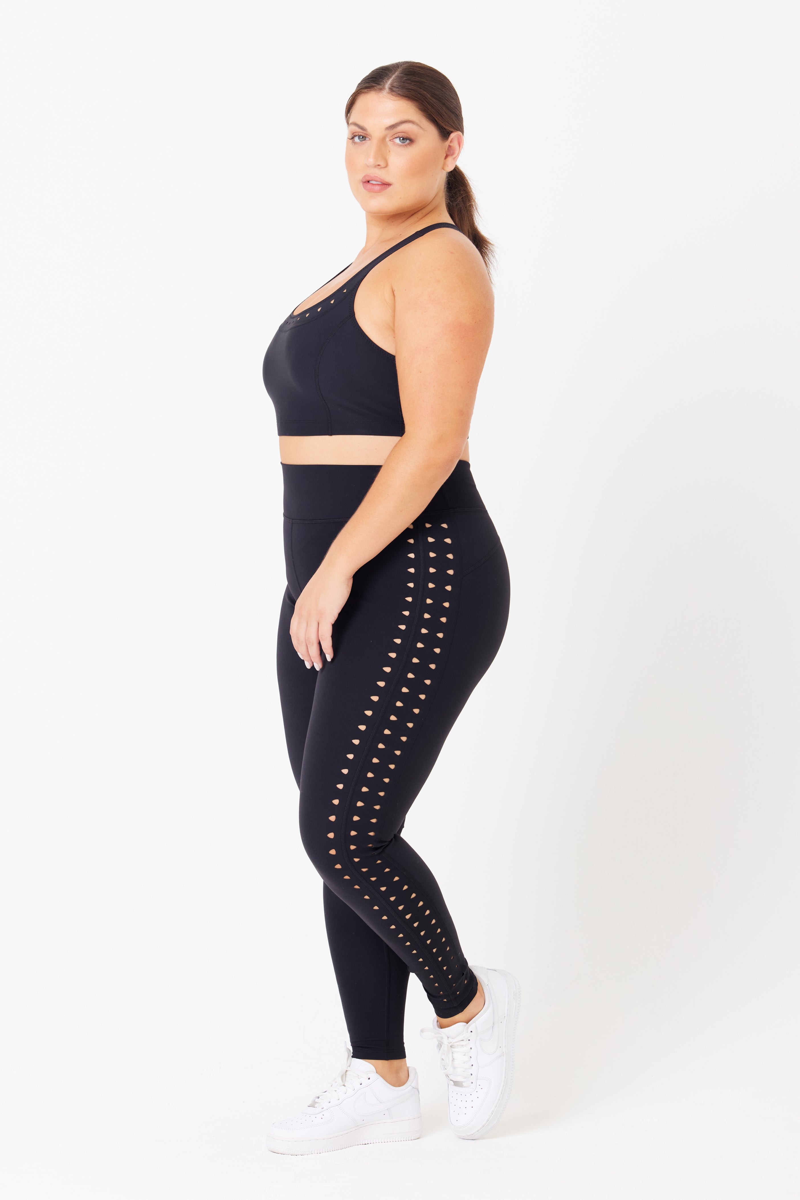 Confidently standing in athletic wear, the woman showcases a Gigi C Sport Annabelle Bra Top made from breathable knit and recycled fabric. She wears black with side cut-outs, pairs it with white sneakers, and sports a sleek ponytail against a plain white background.