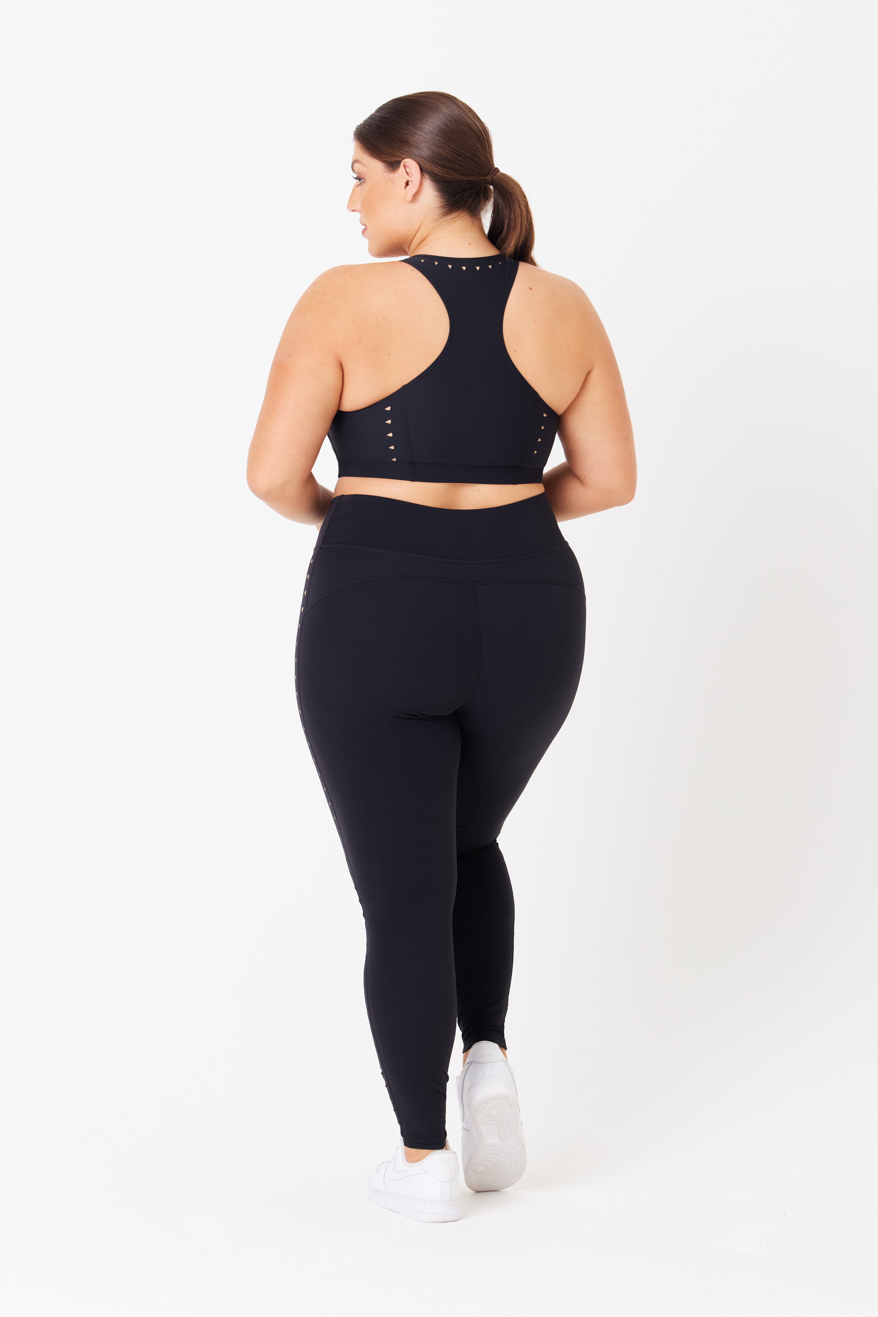 A person in black athletic wear, featuring Gigi C Sports Annabelle Bra Top with cut-out details and recycled fabric leggings, paired with white sneakers. They are facing away from the camera against a plain white backdrop.