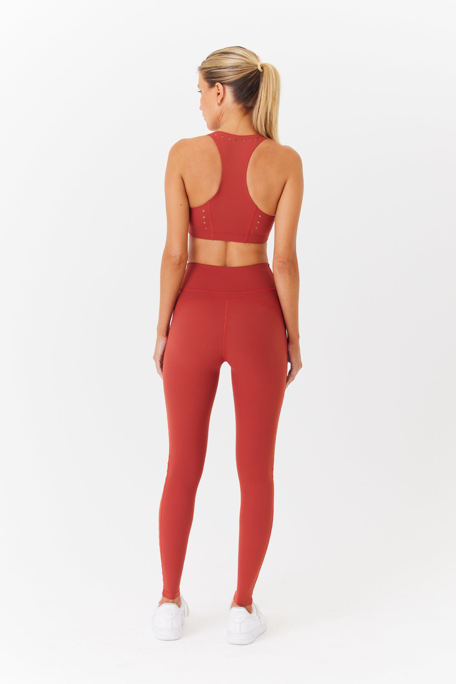 A woman with a ponytail stands on a white background, wearing Gigi C Sports Katrina Legging with sleek laser cutouts and a red sleeveless top. She completes her athletic look with white sneakers.