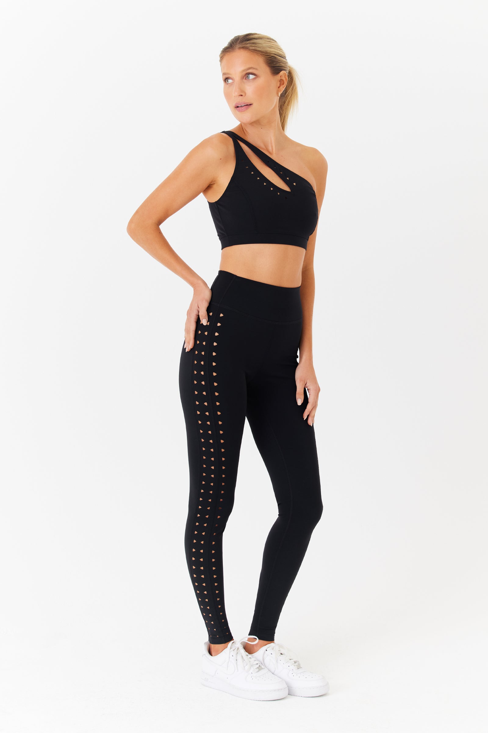 A person confidently poses in a Jacqui Asymmetrical Bra with one-shoulder straps and high-waisted leggings by Gigi C Sport, featuring side perforations. Theyre ready for hot yoga, finishing the look with white sneakers on a plain white background.