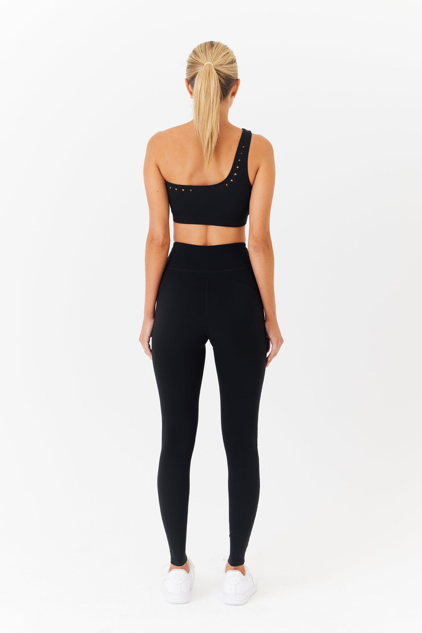Facing away, a person wears a black Jacqui Asymmetrical Bra by Gigi C Sport with high-waisted leggings. Their long blonde hair is in a ponytail, and they wear white sneakers against a plain white background, ready for hot yoga.