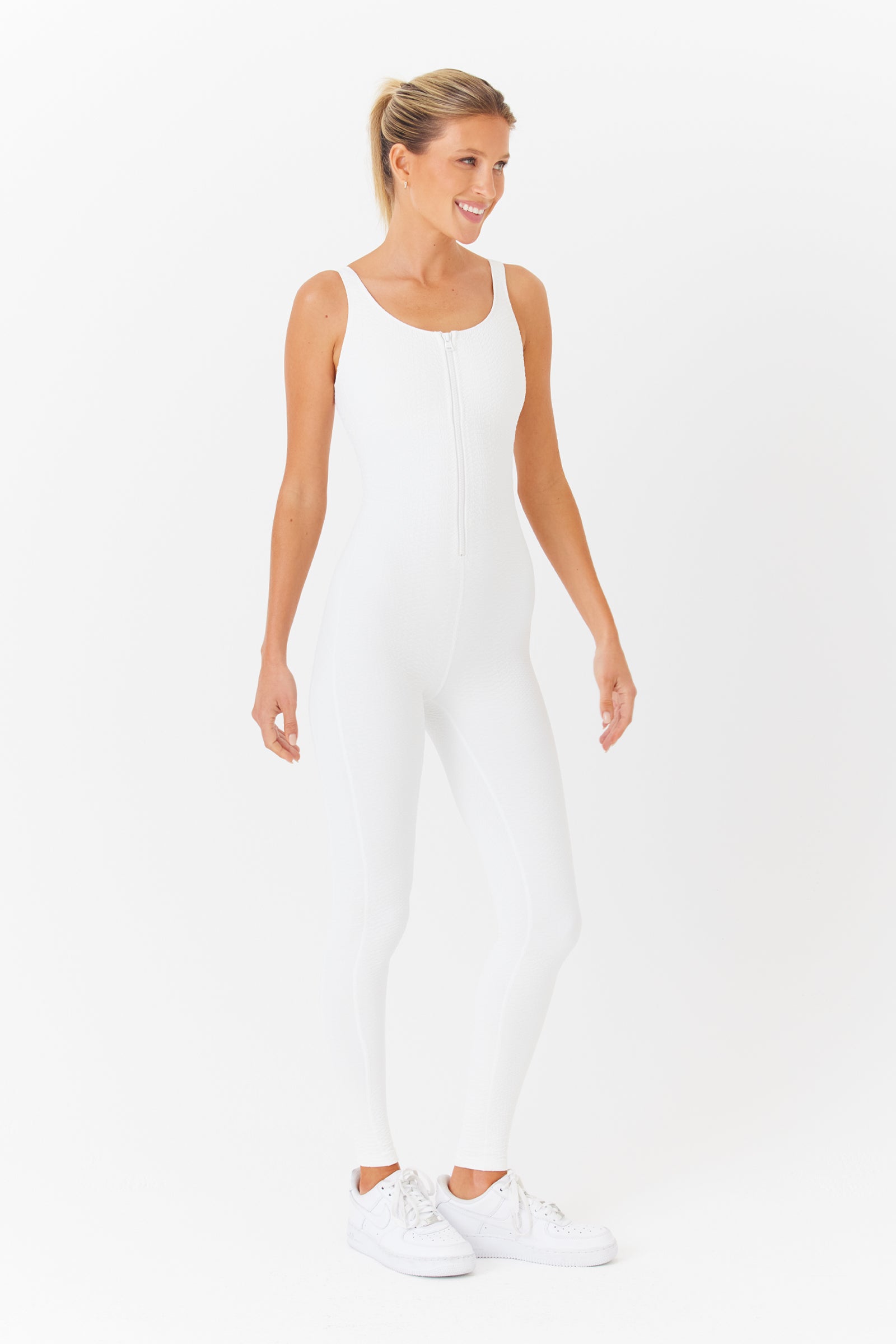 A person poses against a white backdrop wearing the Gigi C Sport Merit Jumpsuit, a sleeveless, zip-up design featuring bi-stretch compression, paired with white sneakers. Their hair is tied back as they smile and angle their body slightly to the side.